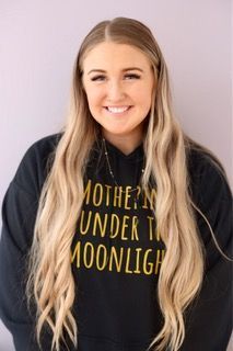 Smiling woman in a black hoodie with gold text against a light purple background