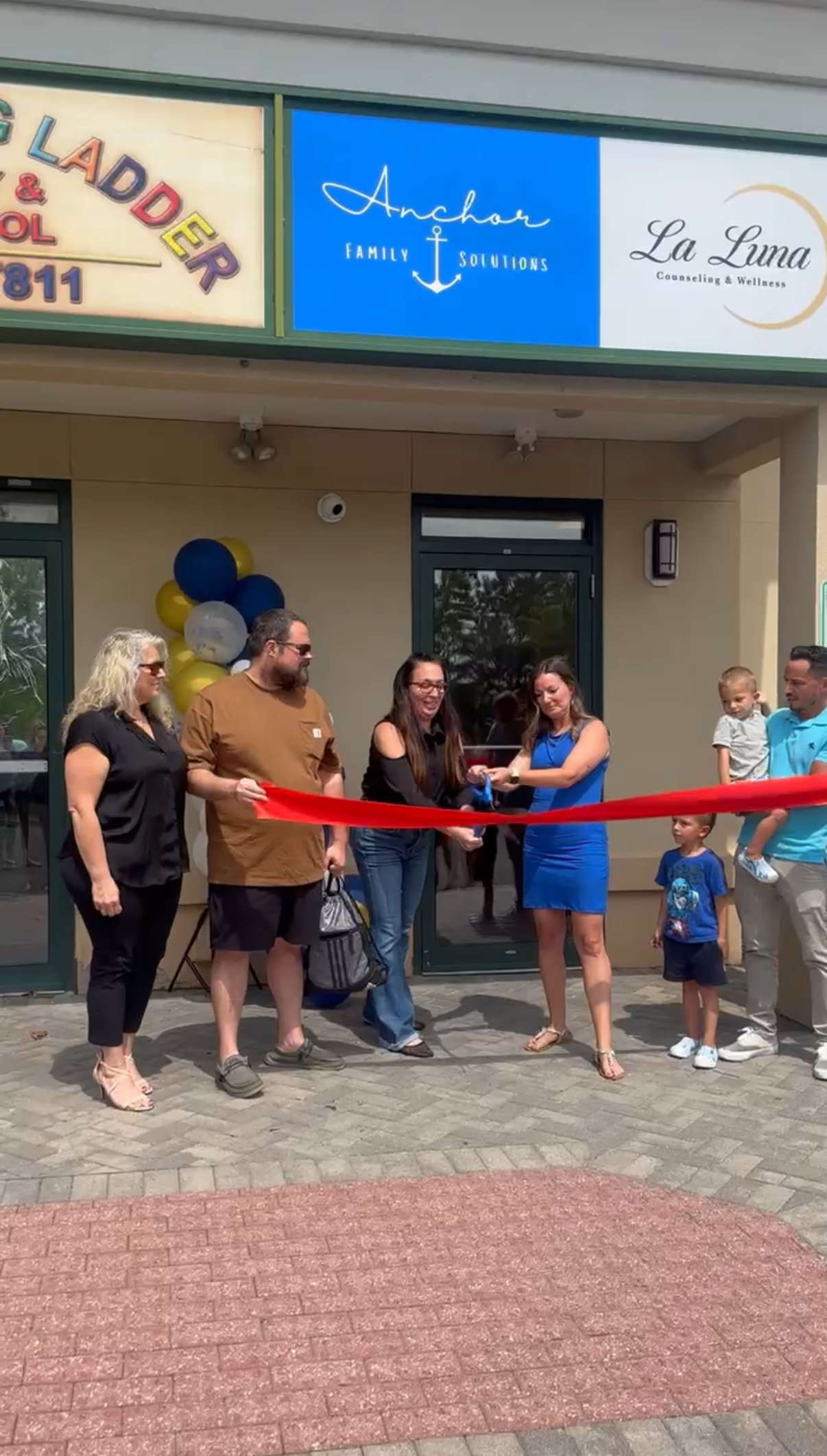 People gathered outside a storefront for a ribbon-cutting ceremony with a red ribbon and balloons.