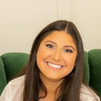 Smiling person with long dark hair seated in a green chair against a light wall