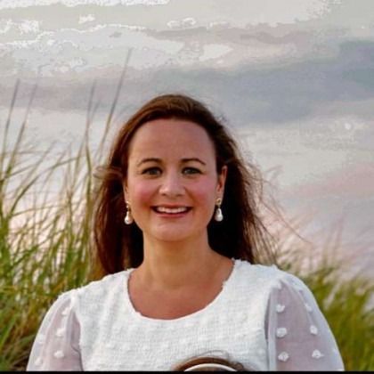 Smiling woman in a white blouse standing outdoors near tall grass and water at sunset