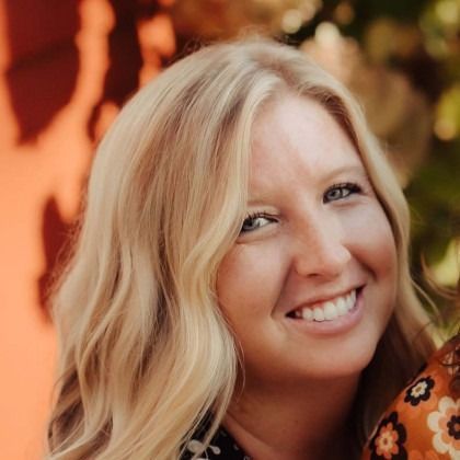 Smiling blonde woman with wavy hair, outdoors with warm orange and green background