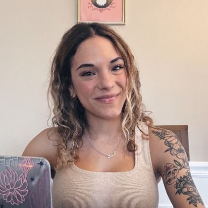 Smiling woman seated indoors, wearing a beige tank top with a floral tattoo on her arm.