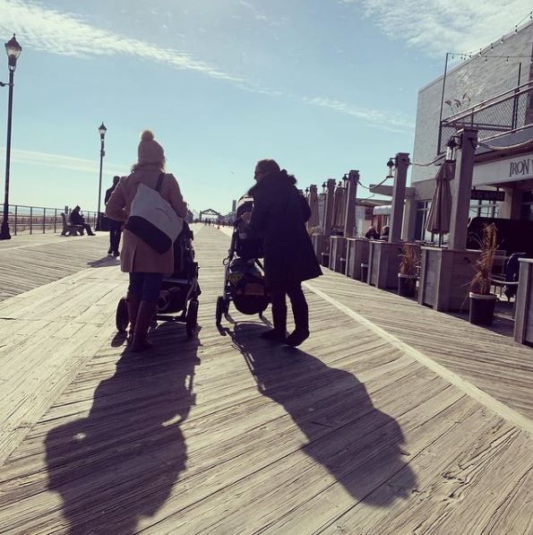 Two people on mobility scooters on a sunny boardwalk by the sea, with long shadows stretching ahead.