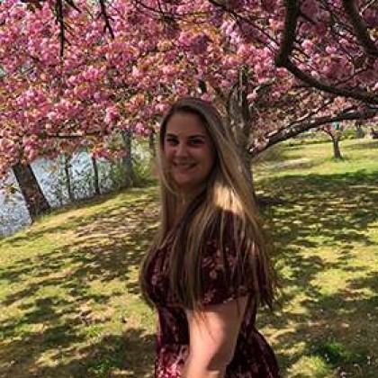 Woman standing under pink blossoms in a sunny park, smiling at the camera.
