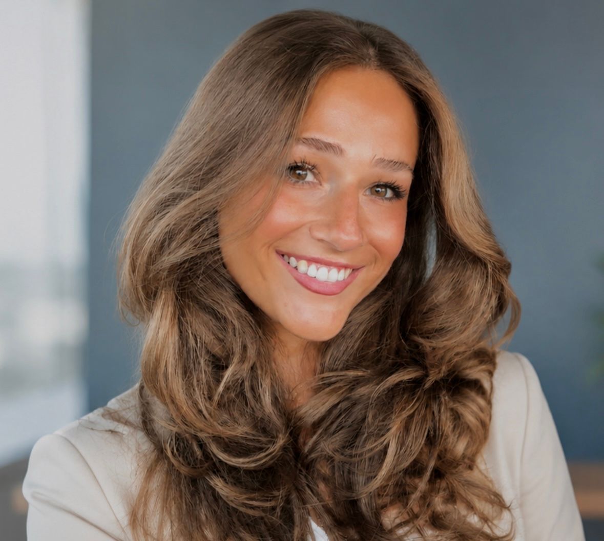 Smiling woman with long wavy brown hair in a light blazer against a blurred gray background