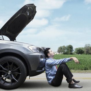 A man is sitting on the ground next to a broken down car with the hood up.