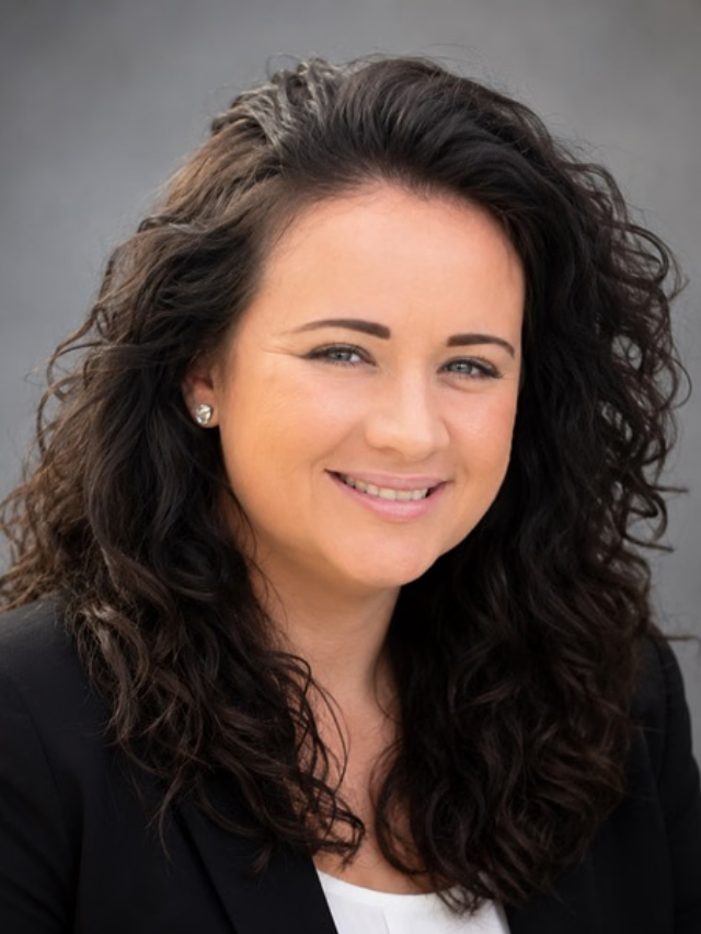 Smiling woman with long dark curly hair, wearing a black blazer against a gray background