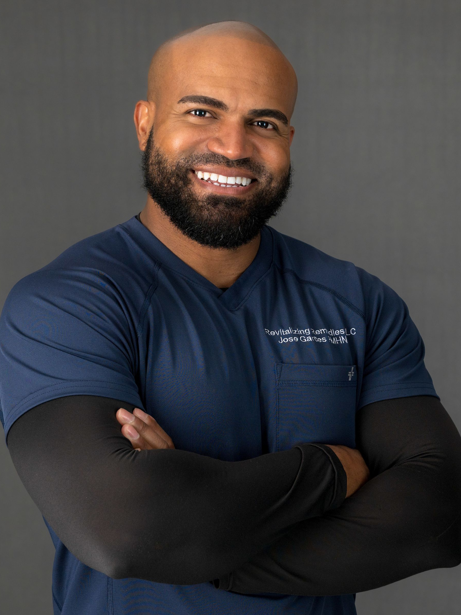 Man with a beard, arms crossed, smiling. Wearing a navy blue shirt with a black undershirt, against a grey backdrop.