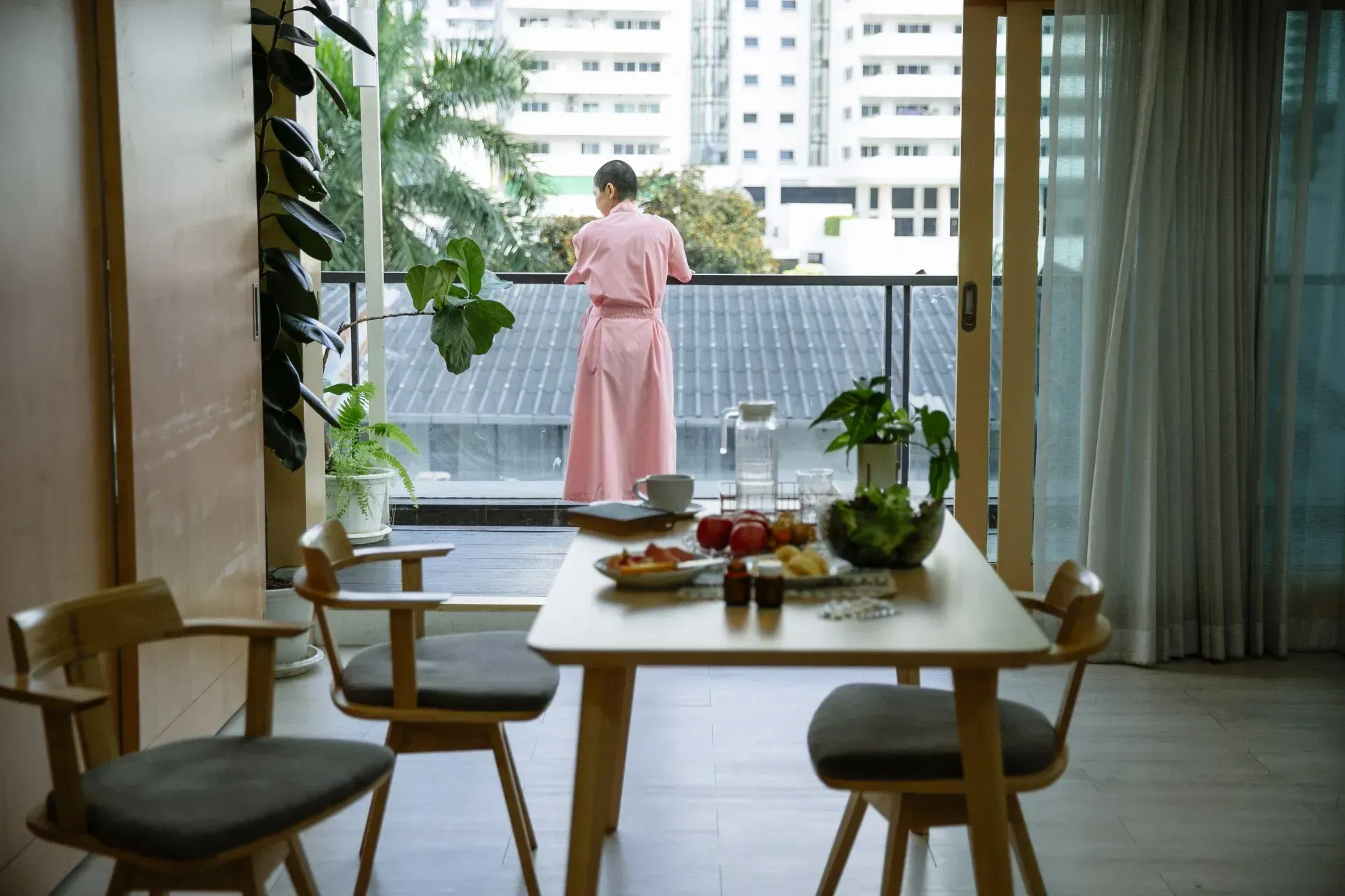 Person in a pink dress stands on a balcony, looking out at buildings beyond a dining table and chairs.