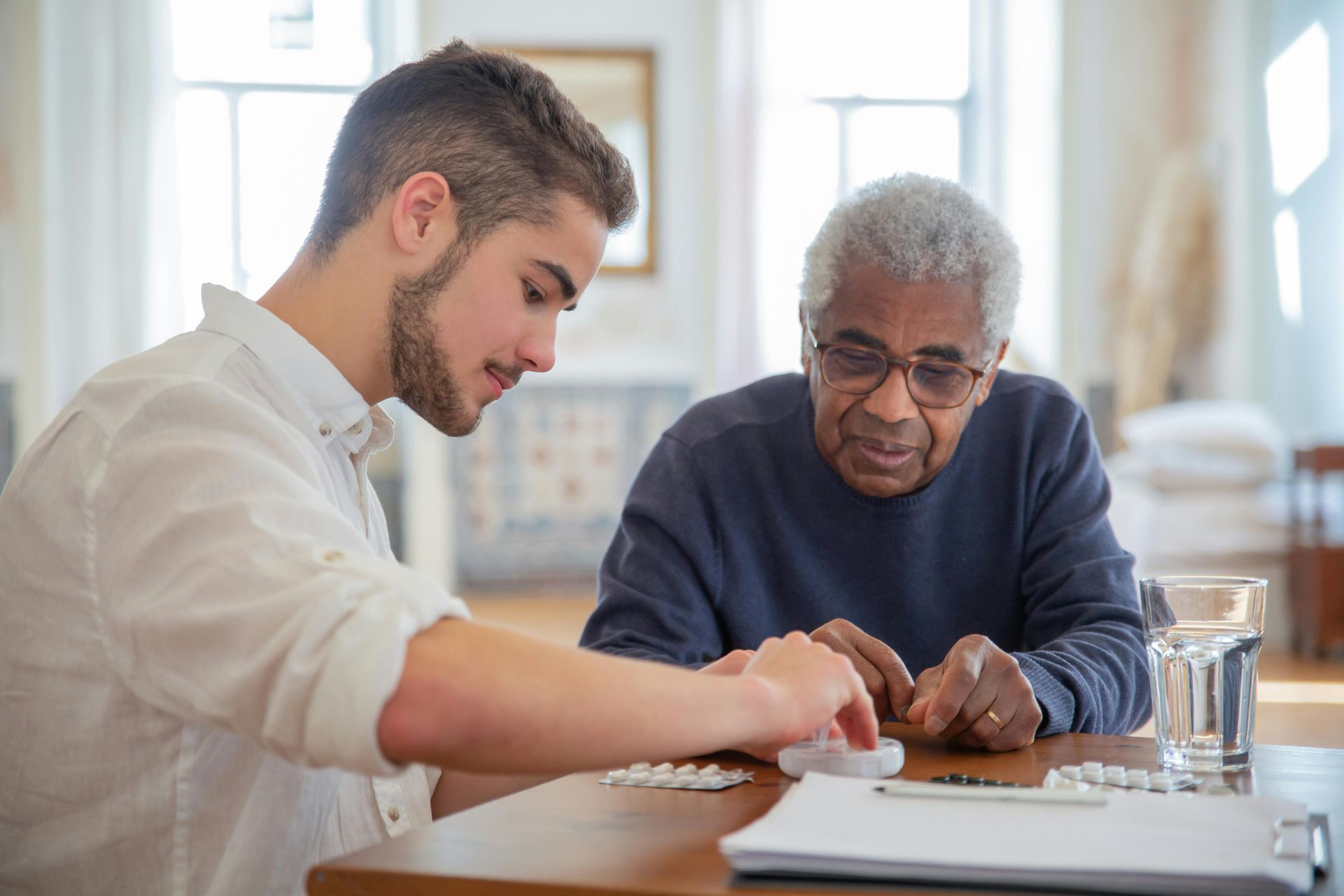 Young man assisting older person with medication at a table. Bright room.