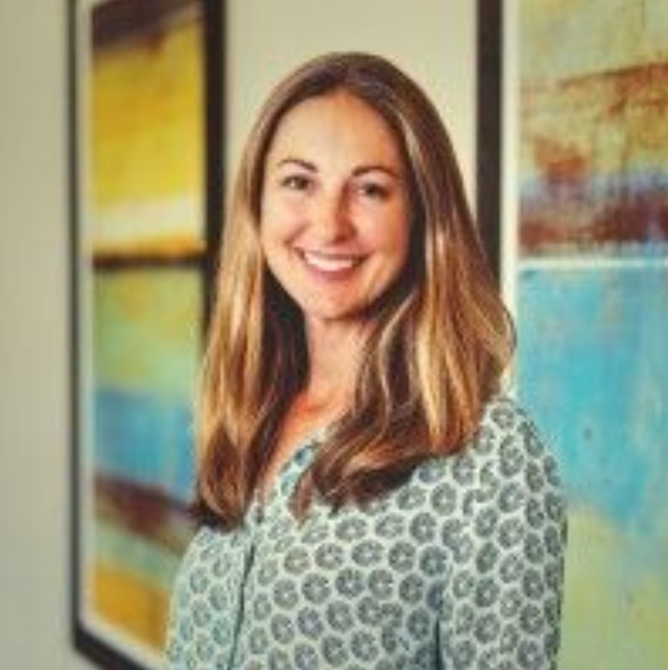 A woman is smiling in front of a painting on a wall.