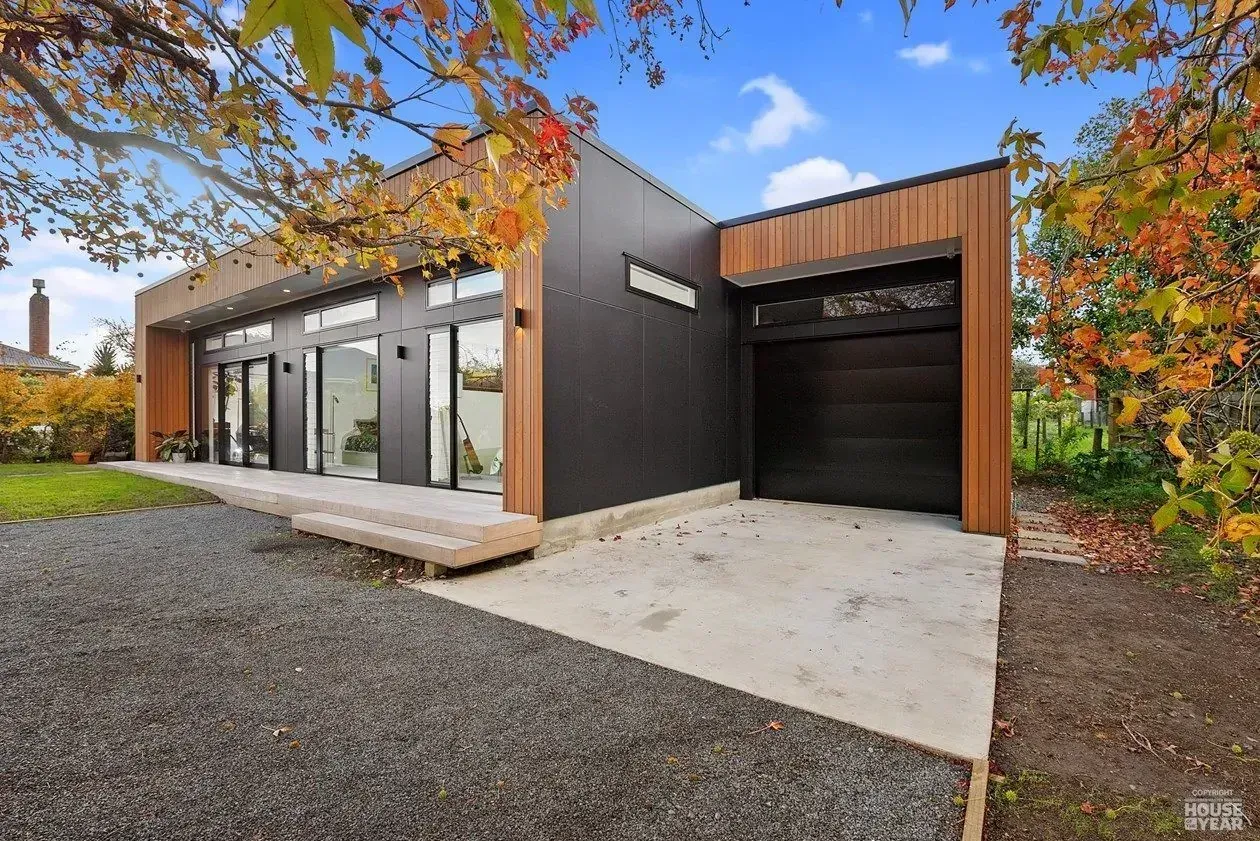 Modern black house with large windows, wood accents, and a garage, set on a gravel drive with autumn trees.