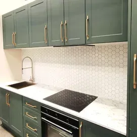 Green kitchen cabinets with brass handles, white countertop, black sink and cooktop, and hex tile backsplash