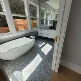 Bright modern bathroom with a freestanding white tub, large windows, and sunlight on gray floor.