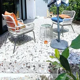 Patio with two wicker chairs, a small glass table, and a gray umbrella base on speckled tile flooring