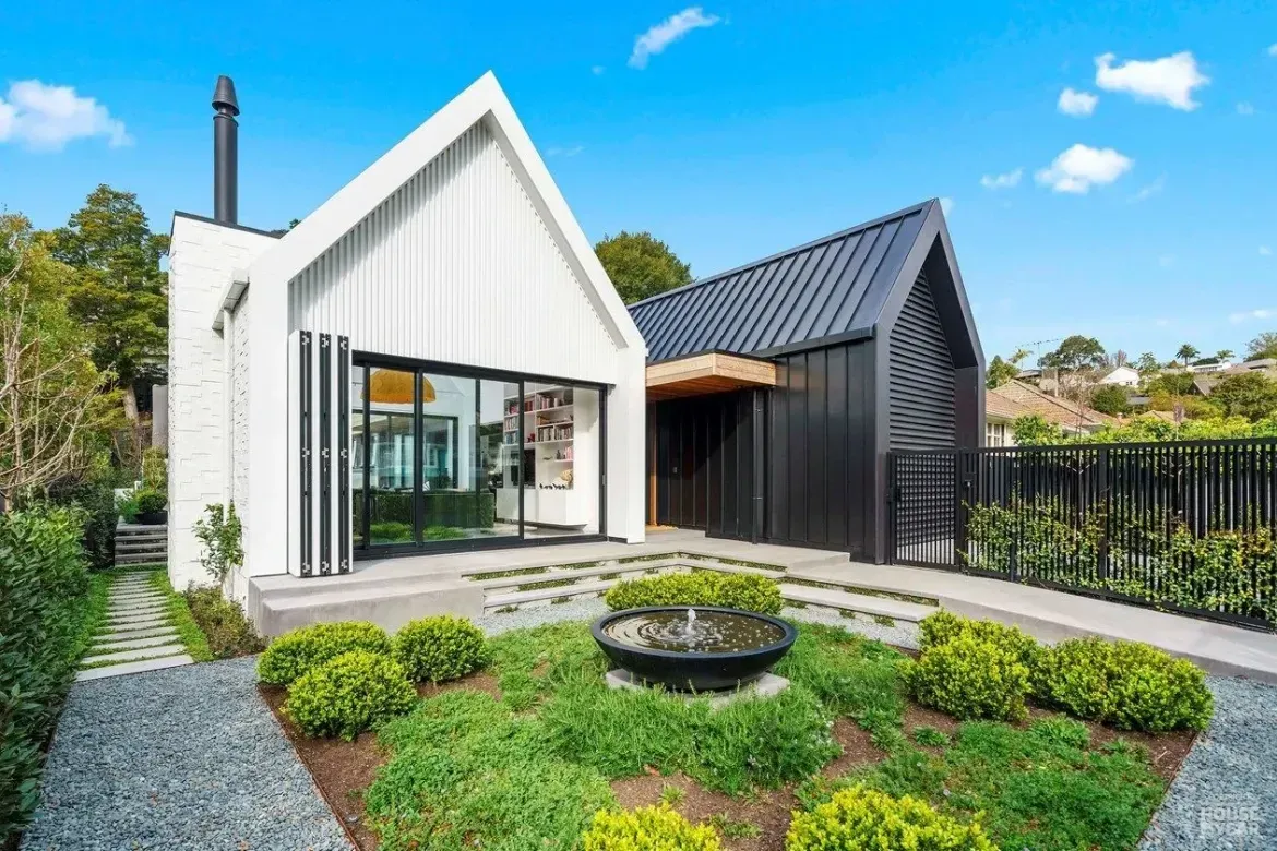 Modern white house with black roof, landscaped front yard, and circular garden feature under a blue sky