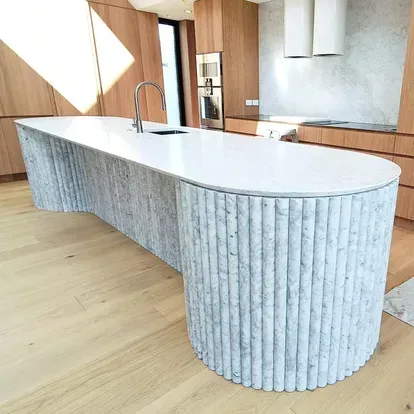 Modern kitchen with a white waterfall island, fluted stone base, wood cabinetry, and light wood floors