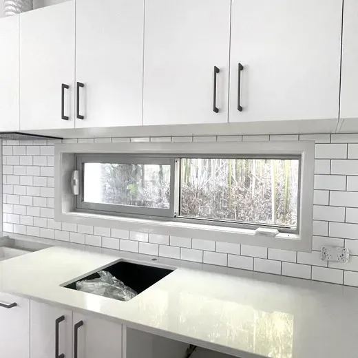 Modern white kitchen with subway tile backsplash, long window, and white cabinets above a countertop sink.