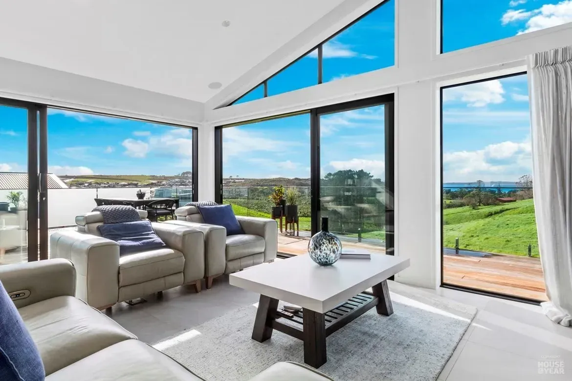 Bright modern living room with large windows, white furniture, and a view of green fields and blue sky.