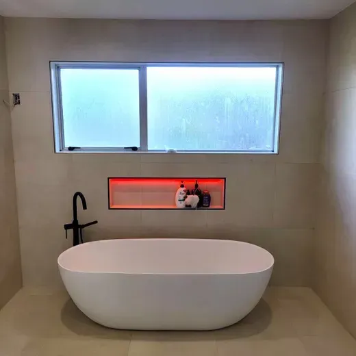Modern bathroom with freestanding tub, frosted window, and lit recessed shelf holding decor