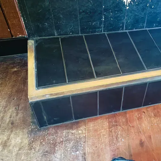 Black tiled step with tan trim beside a brown wooden floor and dark wall panels