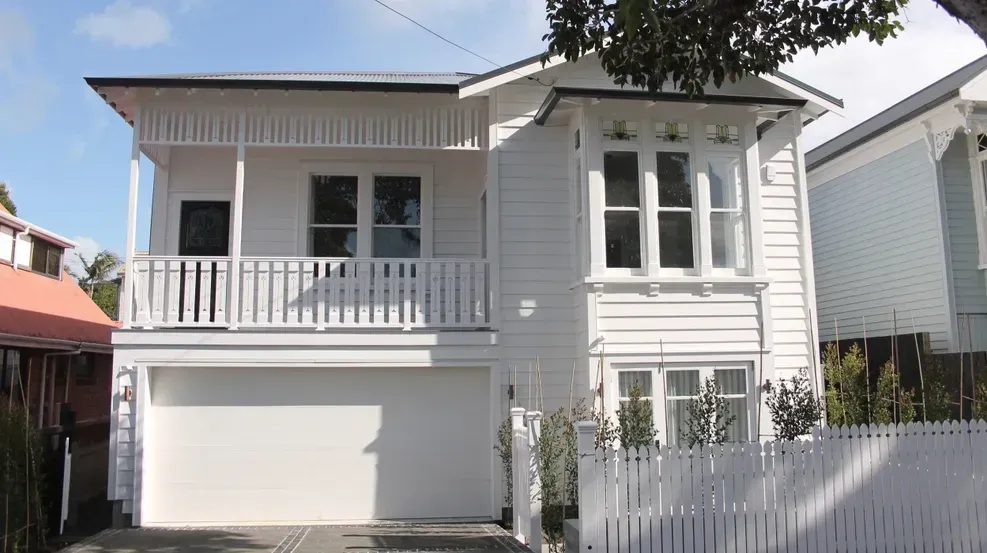 White two-story house with balcony, garage, and white picket fence in a sunny neighborhood