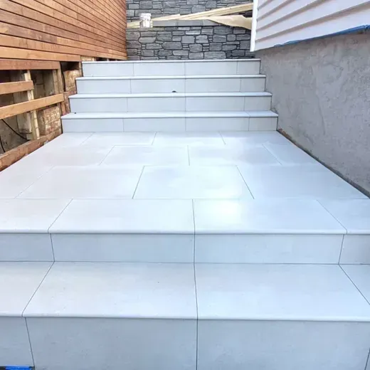 White tiled outdoor steps leading to a small stone landing between a wooden fence and gray wall