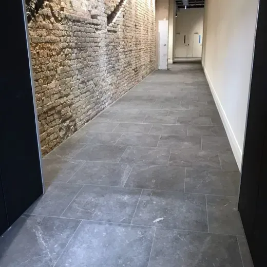 Narrow hallway with a stone wall on the left and gray tile floor, leading to a lit doorway at the end