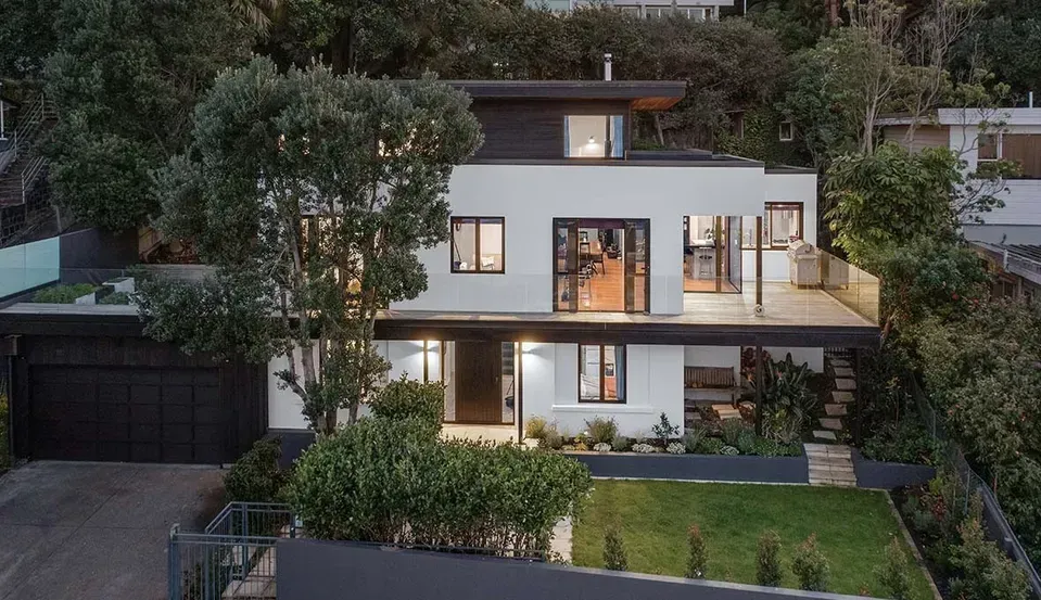 Modern two-story house at dusk with lit windows, lush greenery, and a garage