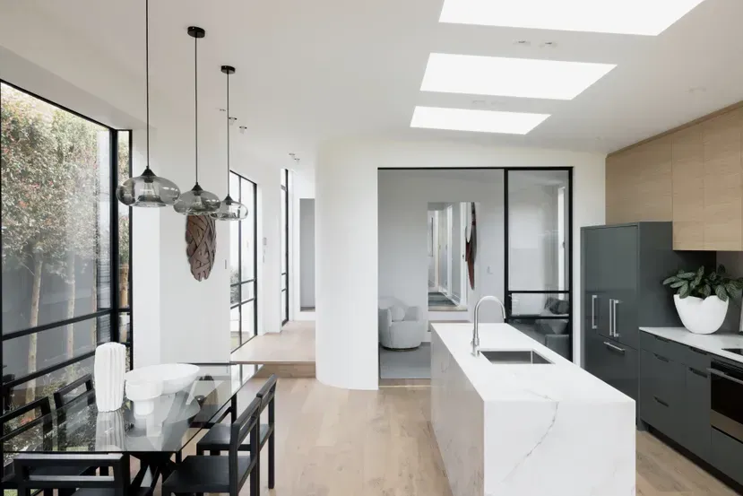 Modern open-plan kitchen and dining area with large windows, white island, black table, and pendant lights