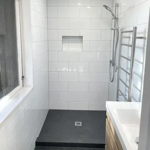 Modern white-tiled accessible shower with dark floor, window, built-in niche, and chrome grab bar