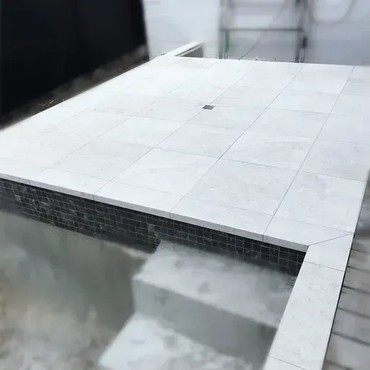 White-tiled pool deck with a small empty pool and steps, bordered by dark mosaic tile edging