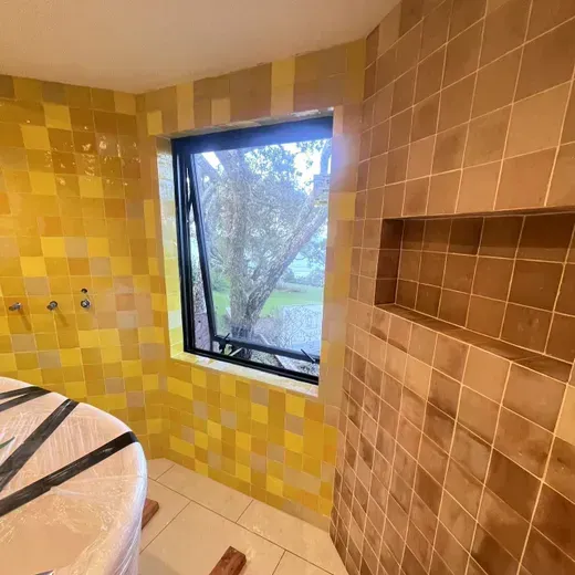 Small tiled bathroom with a window, yellow and beige walls, and a round object at left.