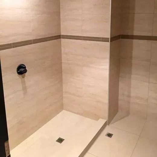 Empty beige tiled shower stall with two floor drains and a black shower knob on the wall