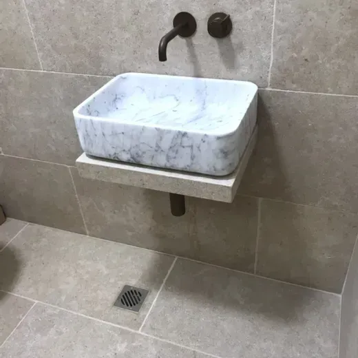 Modern bathroom sink with white marble basin, wall-mounted against beige tiled walls and floor.