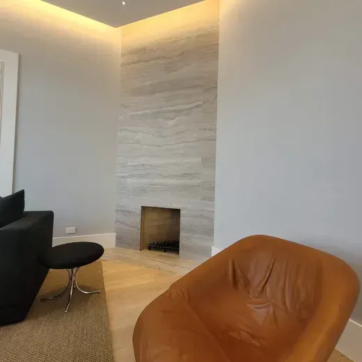 Modern living room corner with a white stone fireplace, tan chair, black chair, and warm ceiling lighting