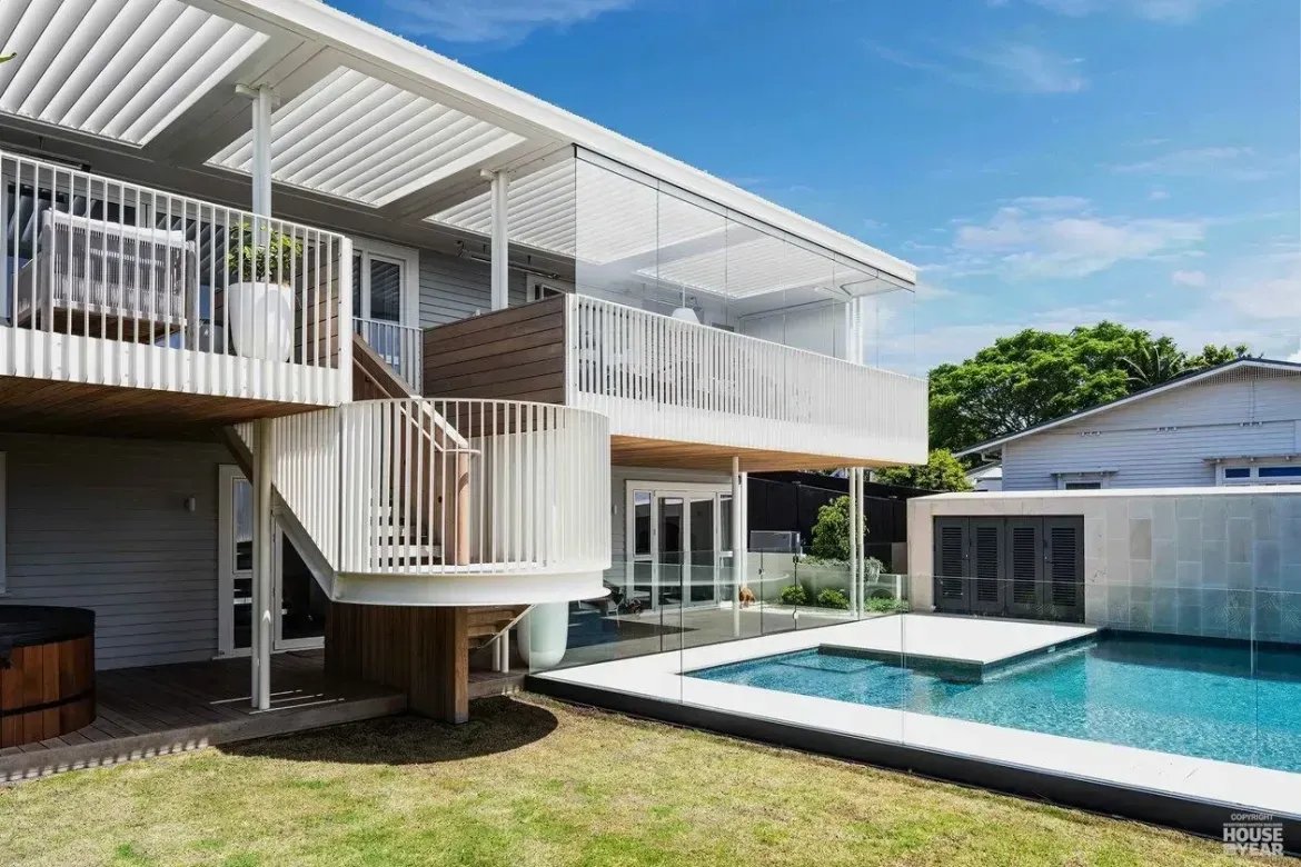 Modern white house with deck and curved staircase beside a backyard swimming pool under a blue sky