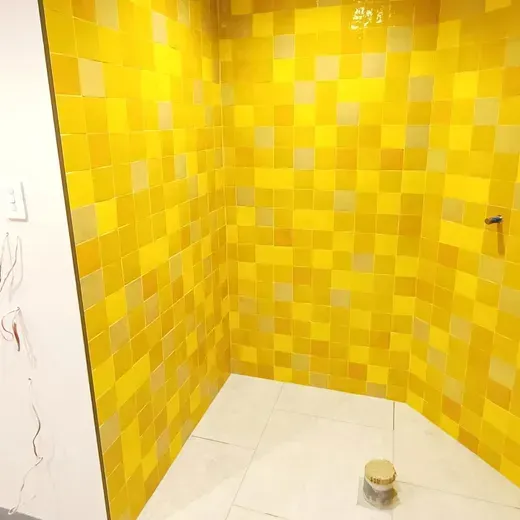 Yellow tiled shower corner with white floor and a small container on the floor