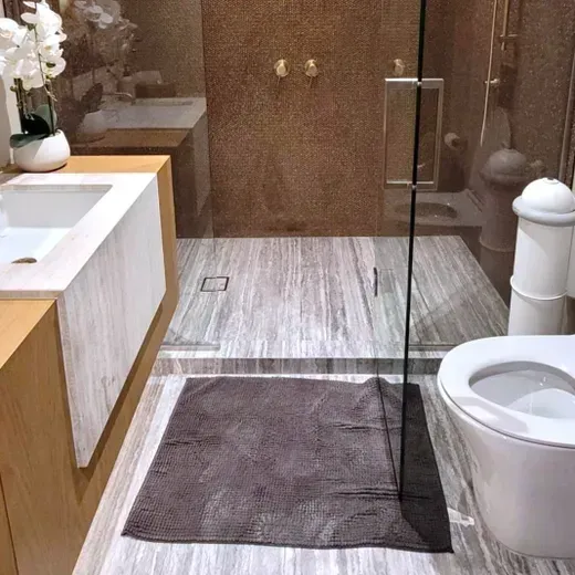 Modern bathroom with white sink, toilet, glass shower, and gray bath mats on a wood-look floor