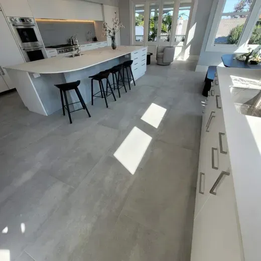 Modern white kitchen with large island, black stools, and bright sunlight on gray floor