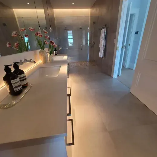 Modern bathroom with double sinks, large mirror, flowers, and a glass shower in a bright hallway