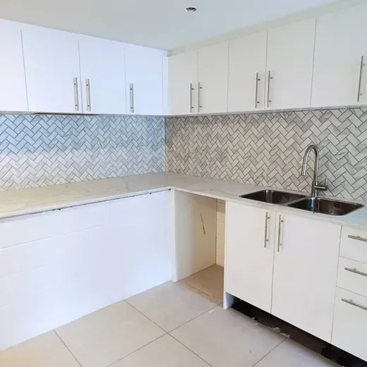 Modern white L-shaped kitchen with white cabinets, gray backsplash, beige tile floor, and double sink