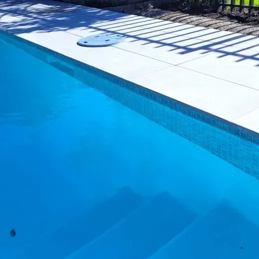 Bright blue swimming pool beside a white tiled deck with ladder shadows and a drain cover