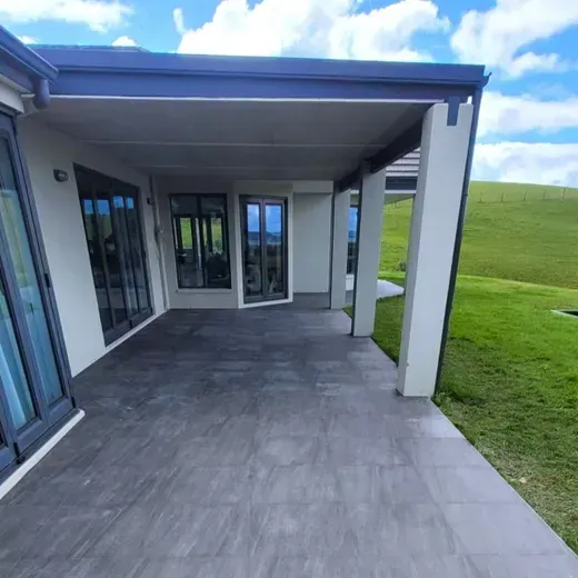 Covered patio with gray pavers, white columns, and glass doors beside a green lawn and hills.