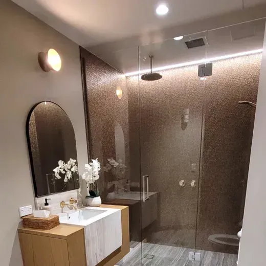 Modern bathroom with glass shower, wooden vanity, oval mirror, and warm wall lighting