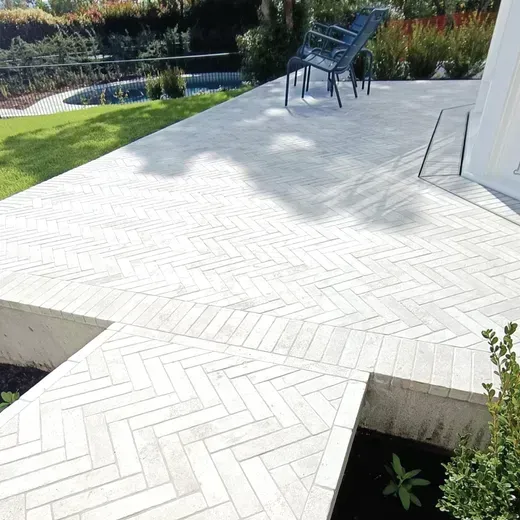 White herringbone patio beside a garden, with chairs and a pool in the background