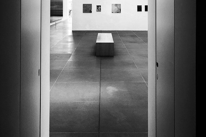 Black-and-white gallery hallway with a bench centered on tiled floor, framed by doorways.