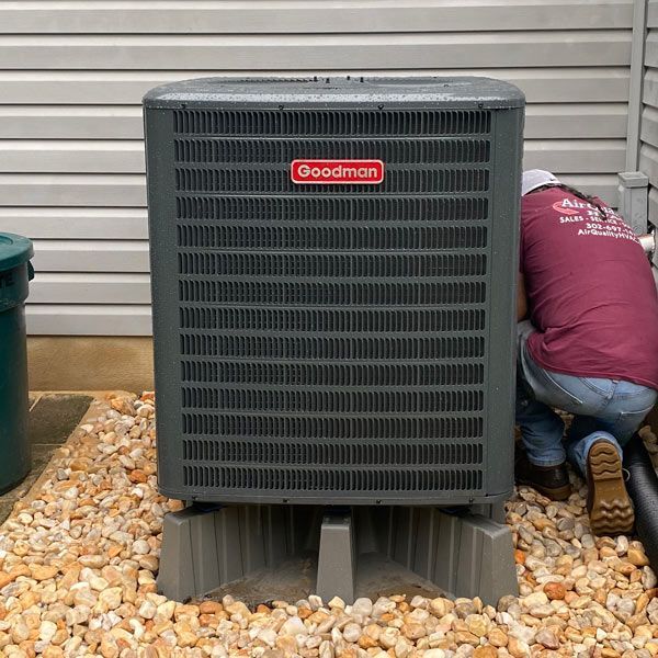 Technician kneels beside a Goodman outdoor HVAC unit against a house wall.