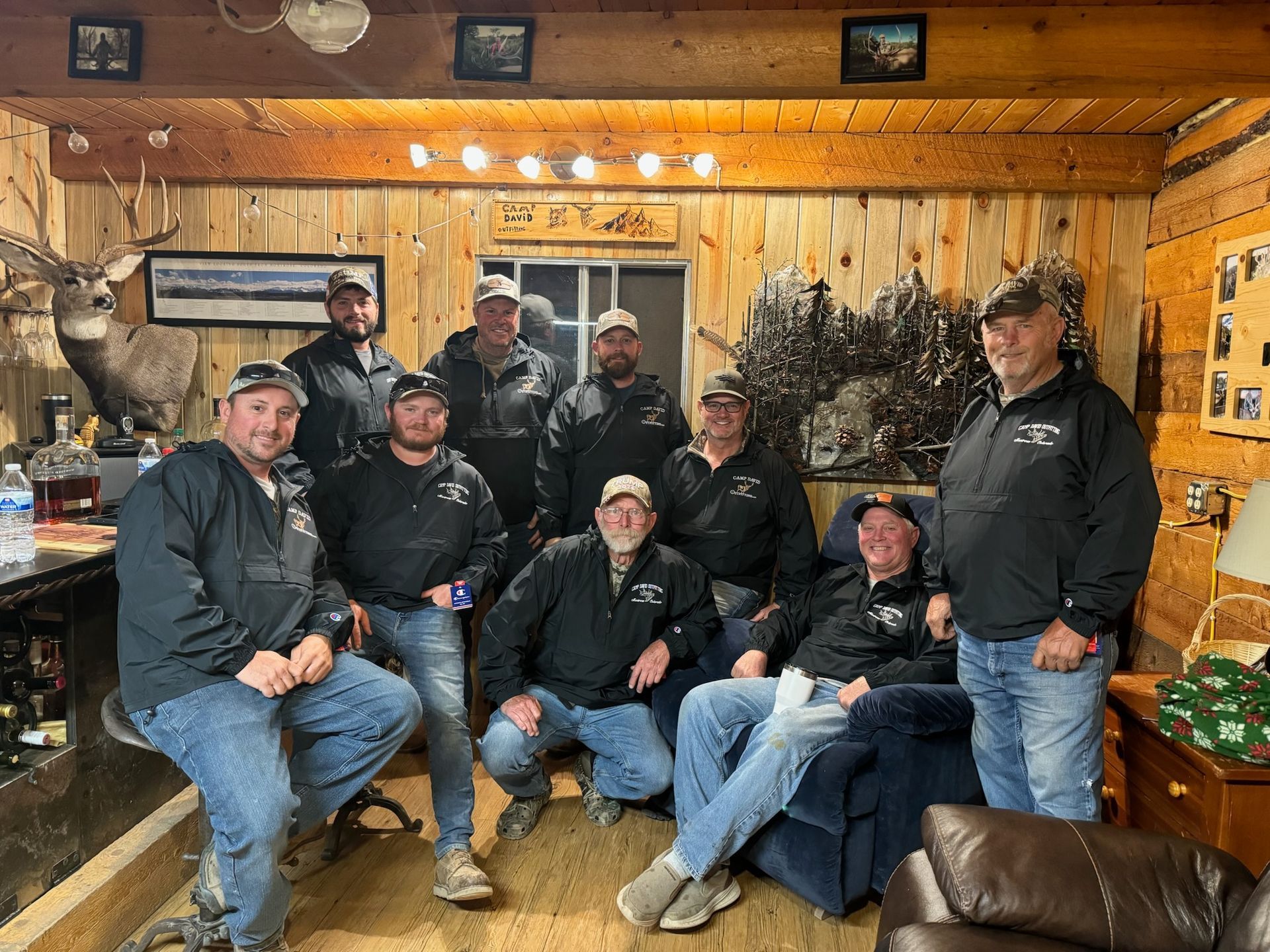 Group of men inside a cabin. Some are seated, others standing. Wood-paneled walls and a deer head are visible.