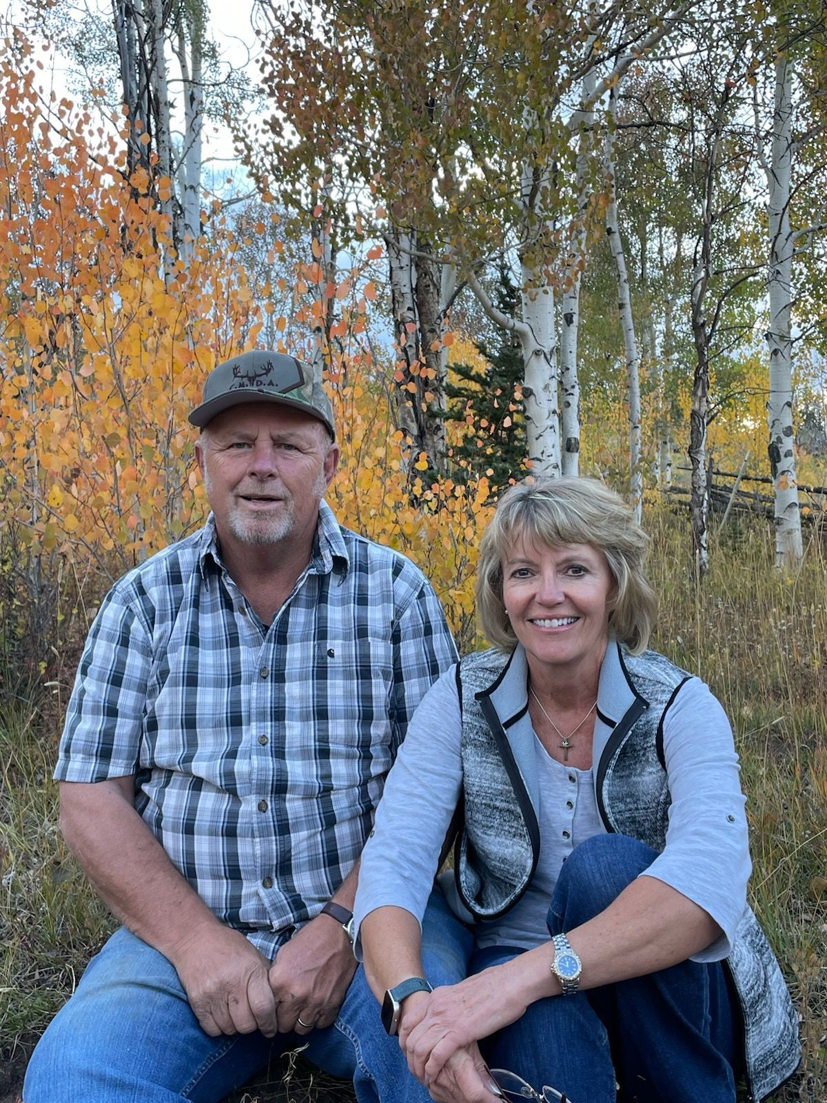 Couple sitting outdoors. Man in plaid shirt, cap; woman in vest. Fall foliage.